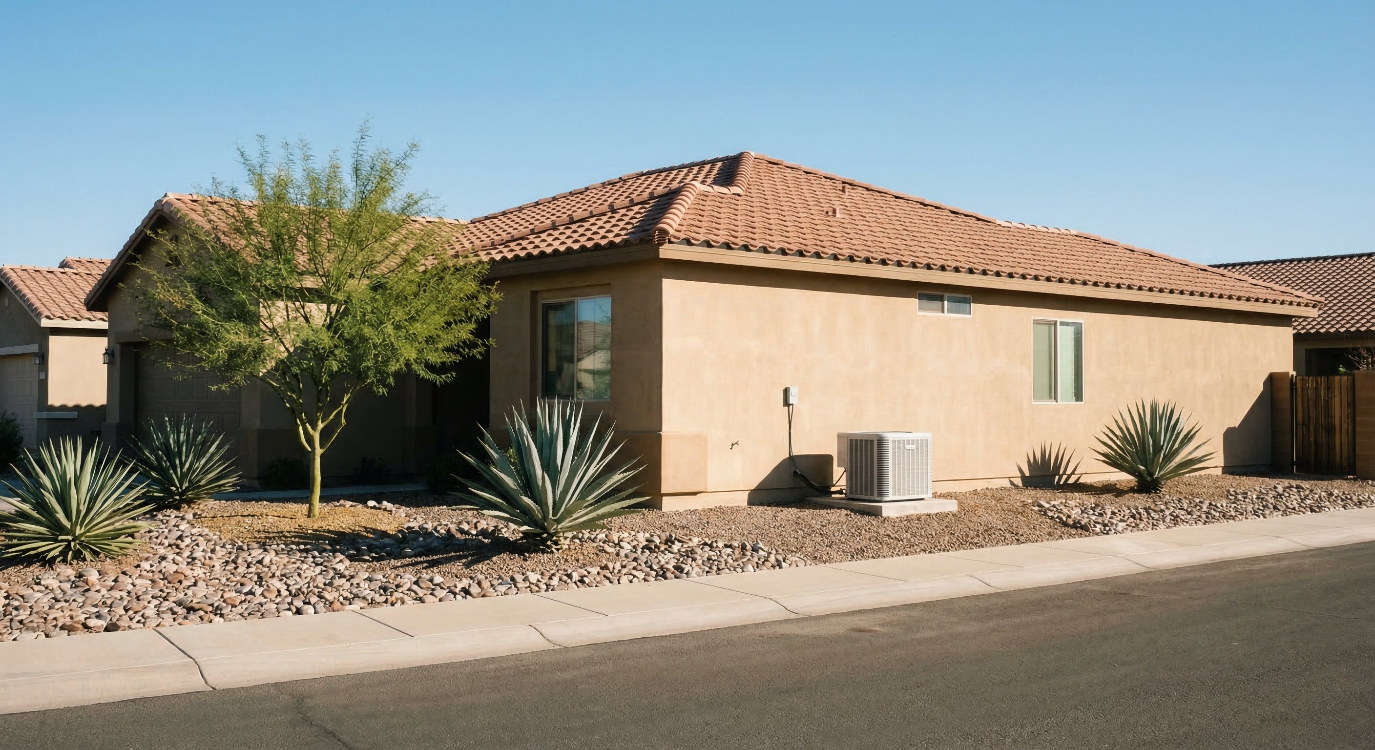 White residential AC condenser unit on concrete pad against tan stucco wall of single-story Phoenix ranch home with desert landscaping, agave plants, river rock, and brilliant blue Arizona sky
