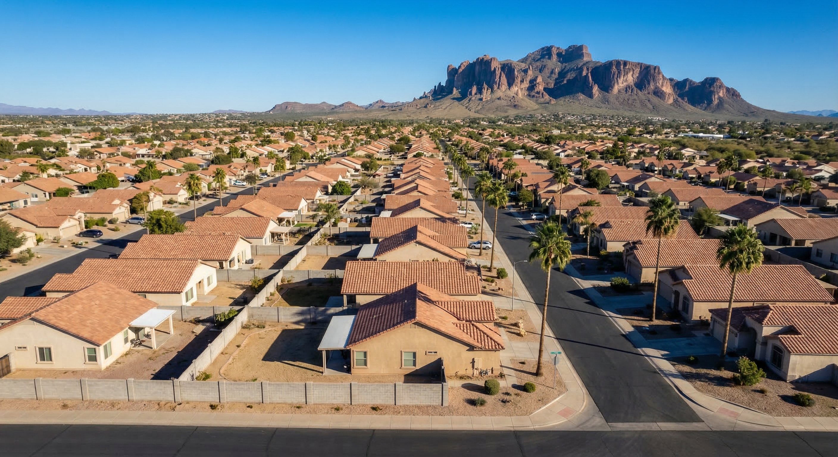 Wide-angle aerial drone view of Apache Junction suburban neighborhood looking east toward the Superstition Mountains, desert landscaping and tile roofs visible under Arizona winter sunshine